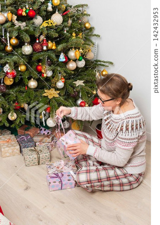A woman aged 52 sits on the floor next to a decorated Christmas tree. She opens gifts on Christmas Day while enjoying the holiday with family members in the background A woman aged 52 sits on the floor next to a decorated Christmas tree. She opens gifts on Christmas Day while enjoying the holiday with family members in the background 142432953