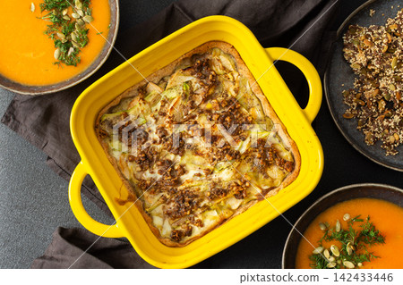 A gluten-free zucchini and walnut pie sits in a yellow dish alongside bowls of pumpkin soup, placed on a dark cloth table. This setup is ideal for a morning or afternoon meal A gluten-free zucchini and walnut pie sits in a yellow dish alongside bowls of pumpkin soup, placed on a dark cloth table. This setup is ideal for a morning or afternoon meal 142433446