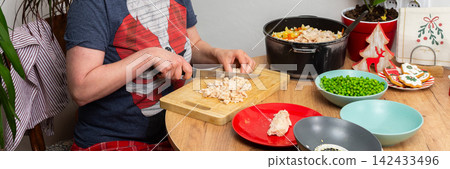 A woman is in her kitchen preparing Olivier salad, Russian salad, for a Christmas and New Year celebration. She is slicing boiled meat on a cutting board with several bowls of ingredients on the table A woman is in her kitchen preparing Olivier salad, Russian salad, for a Christmas and New Year celebration. She is slicing boiled meat on a cutting board with several bowls of ingredients on the table 142433496