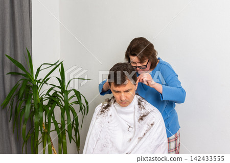 A woman is cutting her husband's hair at home. They are in their living room with plants around. It is a casual afternoon, and he sits in a chair covered with a cape A woman is cutting her husband's hair at home. They are in their living room with plants around. It is a casual afternoon, and he sits in a chair covered with a cape 142433555