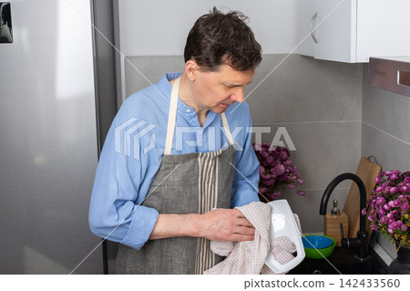 A man stands in the kitchen wiping clean dishes with a cloth. He wears an apron and focuses on his task. The kitchen has a sink and some dishes on the counter A man stands in the kitchen wiping clean dishes with a cloth. He wears an apron and focuses on his task. The kitchen has a sink and some dishes on the counter 142433560