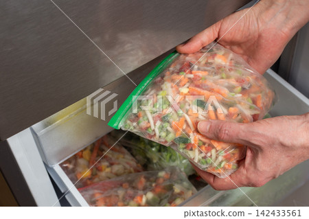 A man reaches into the refrigerator and takes out a bag of frozen vegetables. He is preparing to cook during the afternoon in a kitchen setting A man reaches into the refrigerator and takes out a bag of frozen vegetables. He is preparing to cook during the afternoon in a kitchen setting 142433561