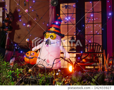 A white inflatable ghost wearing a striped witch hat stands beside a pumpkin on a residential porch. Purple lights, spiders, webs, and eyeballs create a colorful Halloween display at night. A white inflatable ghost wearing a striped witch hat stands beside a pumpkin on a residential porch. Purple lights, spiders, webs, and eyeballs create a colorful Halloween display at night. 142433927