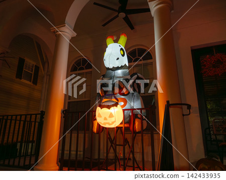 A large inflatable skeleton zebra holds a glowing pumpkin lantern on a decorated house porch. Orange lights illuminate the columns and create a spooky Halloween atmosphere at night. A large inflatable skeleton zebra holds a glowing pumpkin lantern on a decorated house porch. Orange lights illuminate the columns and create a spooky Halloween atmosphere at night. 142433935