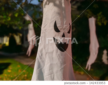 A white hanging ghost decoration sways from a tree during Halloween night. The spooky face creates a festive and eerie neighborhood display. A white hanging ghost decoration sways from a tree during Halloween night. The spooky face creates a festive and eerie neighborhood display. 142433953