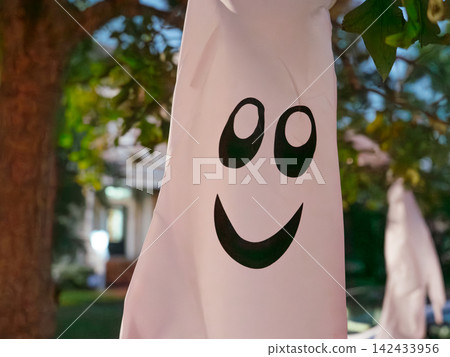 A cheerful ghost decoration hangs from a tree during Halloween evening. The simple white ghost adds playful seasonal charm to an outdoor display. A cheerful ghost decoration hangs from a tree during Halloween evening. The simple white ghost adds playful seasonal charm to an outdoor display. 142433956