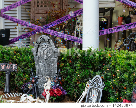 A Halloween yard display features tombstones, skeleton bones, colorful flowers, and haunted caution tape. Floating bubbles add an unexpected playful touch to the spooky outdoor scene. A Halloween yard display features tombstones, skeleton bones, colorful flowers, and haunted caution tape. Floating bubbles add an unexpected playful touch to the spooky outdoor scene. 142433974