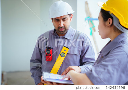 Male and female builders with hard hats and spirit level checking blueprints Male and female builders with hard hats and spirit level checking blueprints 142434696