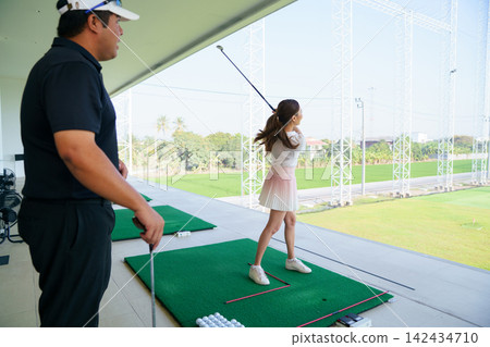 Young Asian woman practicing golf swing with instructor at a driving range Young Asian woman practicing golf swing with instructor at a driving range 142434710