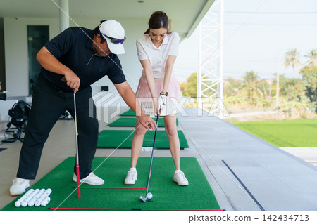 Young Asian woman practicing golf swing with instructor at a driving range Young Asian woman practicing golf swing with instructor at a driving range 142434713