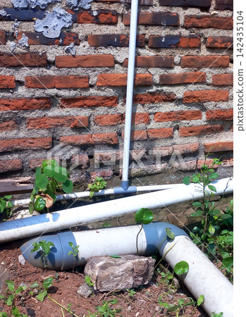 White PVC pipe with elbow joints on the ground, background of a weathered red brick wall. 142435104