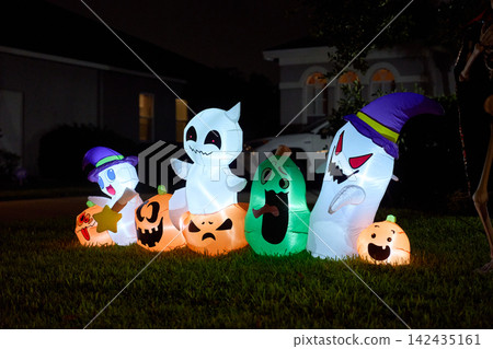 A lineup of smiling ghosts and carved pumpkin inflatables lights a suburban yard. Festive Halloween scene with vivid colors after dark. A lineup of smiling ghosts and carved pumpkin inflatables lights a suburban yard. Festive Halloween scene with vivid colors after dark. 142435161