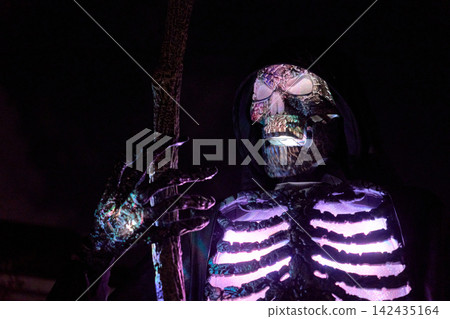 Hooded skull figure with staff shows shimmering LED ribs at night. Multicolor glow lights a spooky Halloween decoration. Hooded skull figure with staff shows shimmering LED ribs at night. Multicolor glow lights a spooky Halloween decoration. 142435164