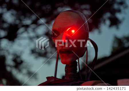 Side profile of an android face with a bright laser-like red eye. Cables and neck tubing add sci-fi detail in a spooky Halloween yard display. Side profile of an android face with a bright laser-like red eye. Cables and neck tubing add sci-fi detail in a spooky Halloween yard display. 142435171