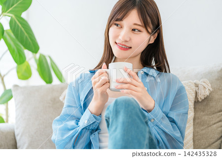 一位女士在家裡的沙發上放鬆,一邊啜飲咖啡和熱飲。 一位女士在家裡的沙發上放鬆,一邊啜飲咖啡和熱飲。 142435328