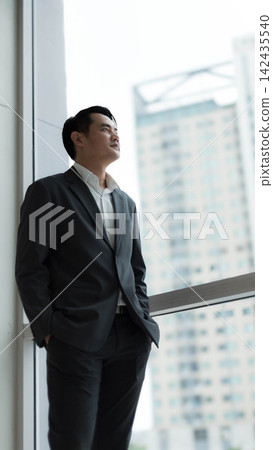Confident Asian businessman in suit standing near glass window looking out at cityscape. Professional corporate executive thinking about future success in modern high rise office skyscraper Confident Asian businessman in suit standing near glass window looking out at cityscape. Professional corporate executive thinking about future success in modern high rise office skyscraper 142435540