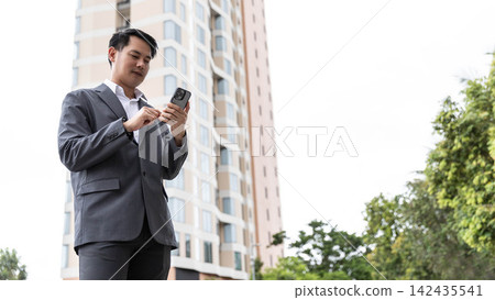 Confident Asian businessman using smartphone outdoors with modern building background. Professional corporate executive checking mobile phone data during work commute in urban residential area Confident Asian businessman using smartphone outdoors with modern building background. Professional corporate executive checking mobile phone data during work commute in urban residential area 142435541