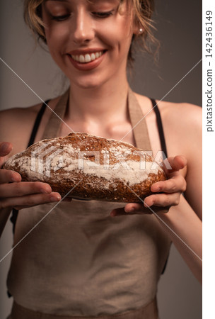 Smiling woman wearing an apron presenting a freshly baked artisan sourdough bread loaf with a crusty texture Smiling woman wearing an apron presenting a freshly baked artisan sourdough bread loaf with a crusty texture 142436149