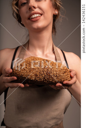 Smiling woman baker presenting artisanal whole wheat bread covered in sesame seeds and wearing a coffee-themed apron Smiling woman baker presenting artisanal whole wheat bread covered in sesame seeds and wearing a coffee-themed apron 142436151