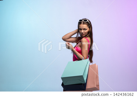 Young woman enjoying successful shopping trip, carrying colorful bags, sunglasses on head, looking at camera on pastel background Young woman enjoying successful shopping trip, carrying colorful bags, sunglasses on head, looking at camera on pastel background 142436162