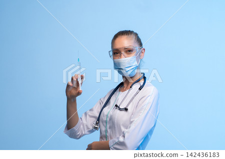 Healthcare worker wearing a protective face mask and safety glasses, holding a syringe with a clear liquid 142436183