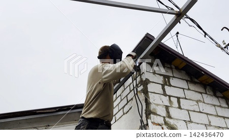 Manual laborer in protective mask welds iron detail at construction. Professional young repairman with welding machine working at the site. Male welder operating with metal on house. Close up Manual laborer in protective mask welds iron detail at construction. Professional young repairman with welding machine working at the site. Male welder operating with metal on house. Close up 142436470