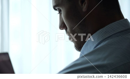 Close up portrait of young entrepreneur standing near large window in modern office and working on notebook. Handsome businessman in shirt using notebook for job with bright day light at background Close up portrait of young entrepreneur standing near large window in modern office and working on notebook. Handsome businessman in shirt using notebook for job with bright day light at background 142436608