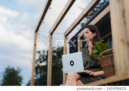 Young woman sitting on tiny house patio, working on laptop. Young woman sitting on tiny house patio, working on laptop. 142436628