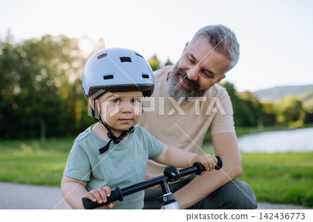 Father helping toddler son learn to ride balance bike for the first time. Father helping toddler son learn to ride balance bike for the first time. 142436773