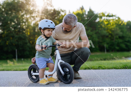 Father teaching toddler son to ride balance bike while wearing safety helmet. 142436775