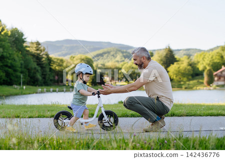 Father teaching toddler son to ride balance bike while wearing safety helmet. Father teaching toddler son to ride balance bike while wearing safety helmet. 142436776