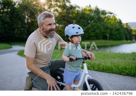 Father helping toddler son learn to ride balance bike for the first time. Father helping toddler son learn to ride balance bike for the first time. 142436796