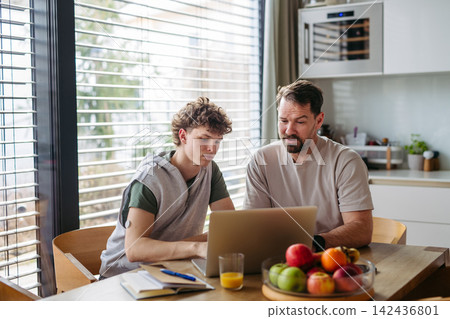 Father helping teenage son with diabetes with homework on laptop in kitchen. Father helping teenage son with diabetes with homework on laptop in kitchen. 142436801