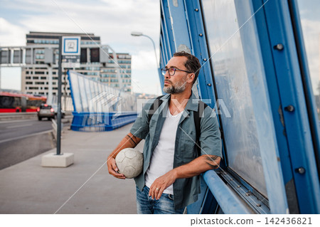 Bearded man with ball under arm using phone while waiting for bus. Bearded man with ball under arm using phone while waiting for bus. 142436821