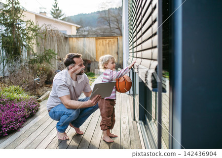 Father controlling smart home exterior blinds using tablet while standing with toddler son. Father controlling smart home exterior blinds using tablet while standing with toddler son. 142436904