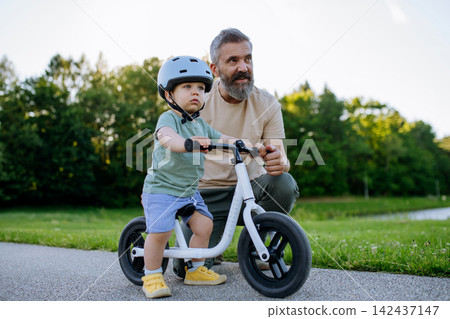 Father teaching toddler son to ride balance bike while wearing safety helmet. 142437147
