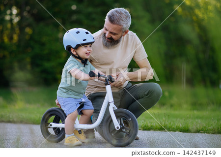 Dad encouraging young child to build confidence on balance bike. 142437149