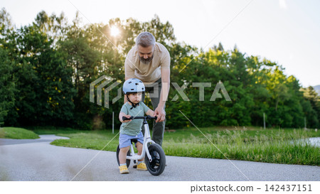 Father teaching toddler son to ride balance bike while wearing safety helmet. Father teaching toddler son to ride balance bike while wearing safety helmet. 142437151