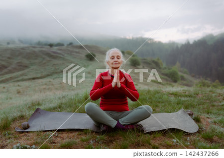 Senior woman meditating in nature on early morning with fog and mountains in background. Senior woman meditating in nature on early morning with fog and mountains in background. 142437266