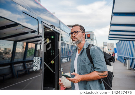 Bearded man with ball under arm using phone while waiting for bus. Bearded man with ball under arm using phone while waiting for bus. 142437476