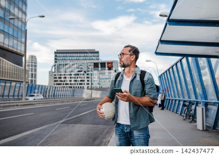 Bearded man with ball under arm using phone while waiting for bus. Bearded man with ball under arm using phone while waiting for bus. 142437477