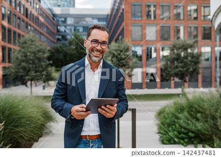 Businessman in front of office building, holding tablet and reading something. Businessman in front of office building, holding tablet and reading something. 142437481