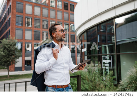 Businessman in front of office building, holding tablet and reading something. Businessman in front of office building, holding tablet and reading something. 142437482