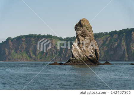 Three Brothers sea stack rises from blue Pacific Ocean with rugged green Kamchatka cliffs behind. Coastal landmark, geology, wilderness tourism, exploration. Three Brothers sea stack rises from blue Pacific Ocean with rugged green Kamchatka cliffs behind. Coastal landmark, geology, wilderness tourism, exploration. 142437505