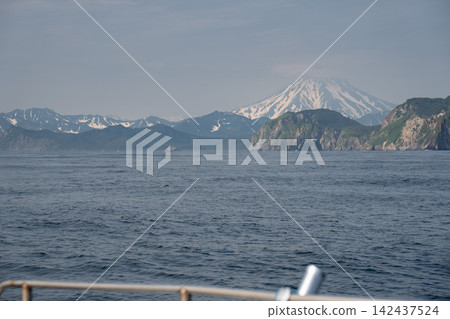 Snow-covered Vilyuchinsky Volcano rises beyond rocky coast and blue sea, viewed from sightseeing catamaran in Kamchatka. Travel, sailing, adventure, nature tourism Snow-covered Vilyuchinsky Volcano rises beyond rocky coast and blue sea, viewed from sightseeing catamaran in Kamchatka. Travel, sailing, adventure, nature tourism 142437524