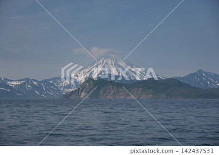 Snowy Vilyuchinsky Volcano rises behind green headland and calm Pacific water in Avacha Bay, Kamchatka, Russia. Nature tourism, volcanic landscape, wilderness, exploration. Snowy Vilyuchinsky Volcano rises behind green headland and calm Pacific water in Avacha Bay, Kamchatka, Russia. Nature tourism, volcanic landscape, wilderness, exploration. 142437531