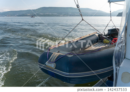 Inflatable motorboat with outboard engine hangs beside moving catamaran with Kamchatka coast in background. Marine transport, boating equipment, navigation, travel, adventure Inflatable motorboat with outboard engine hangs beside moving catamaran with Kamchatka coast in background. Marine transport, boating equipment, navigation, travel, adventure 142437533