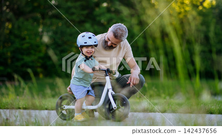 Father teaching toddler son to ride balance bike while wearing safety helmet. 142437656