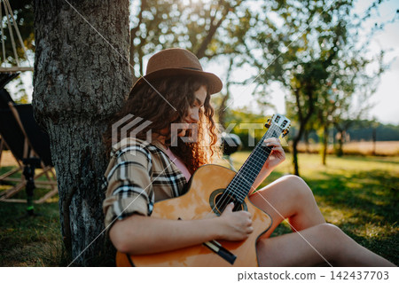 Young beautiful woman is on a camping trip in nature, sitting by tree and playing guitar. Young beautiful woman is on a camping trip in nature, sitting by tree and playing guitar. 142437703