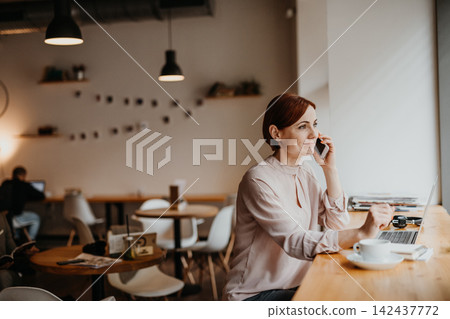 Redhead woman in coffe shop, holding smartphone and making phone reservation. Redhead woman in coffe shop, holding smartphone and making phone reservation. 142437772